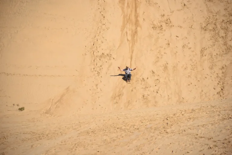 A sandboarder glides down the dunes sitting on a board