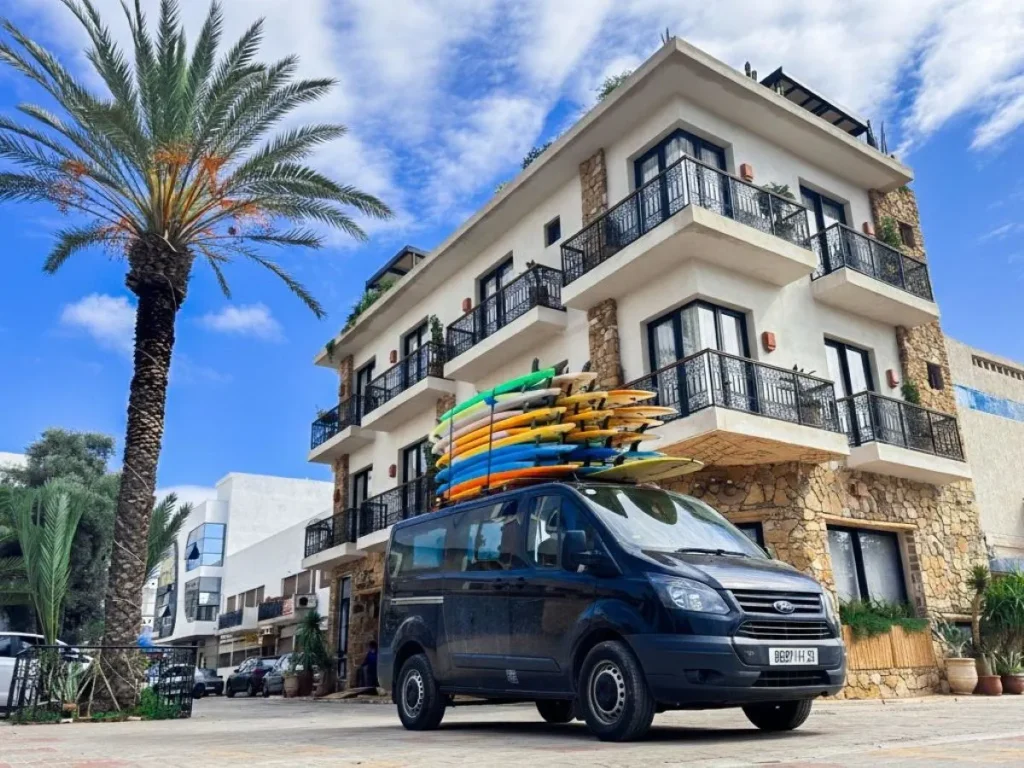 The van is parked in front of the hotel, ready to pick up the surf guests with numerous surfboards on the roof