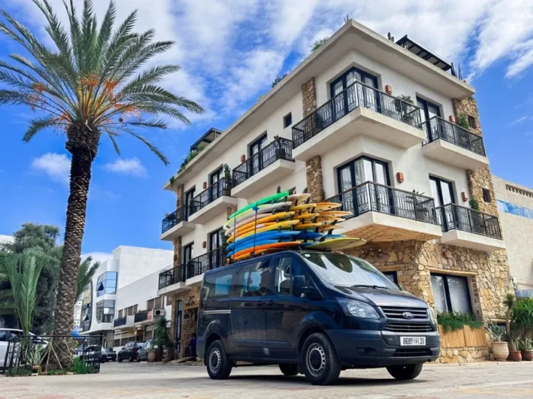 The van is parked in front of the hotel, ready to pick up the surf guests with numerous surfboards on the roof